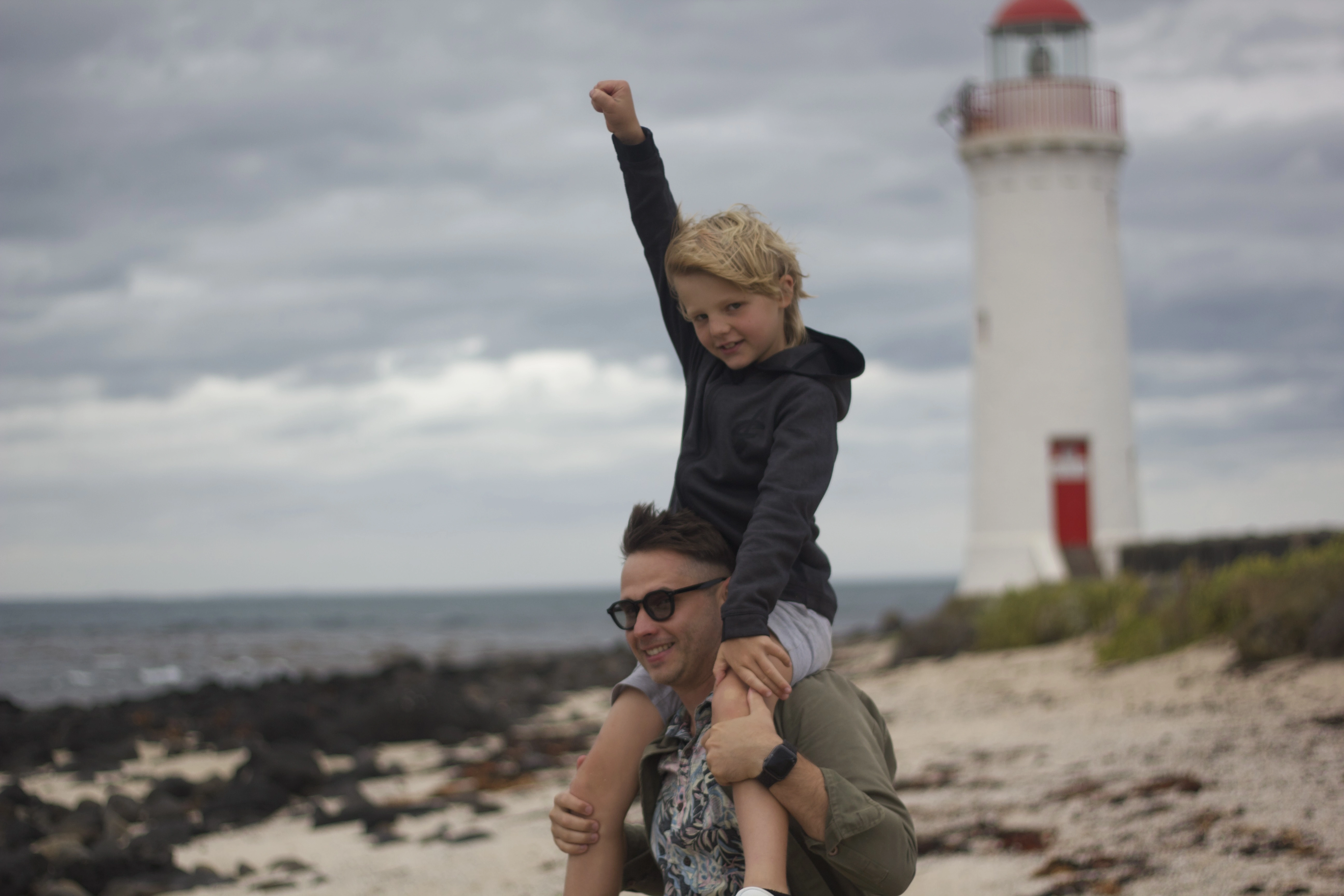 Father carrying young son on his shoulders on an Australian beach, lighthouse in background